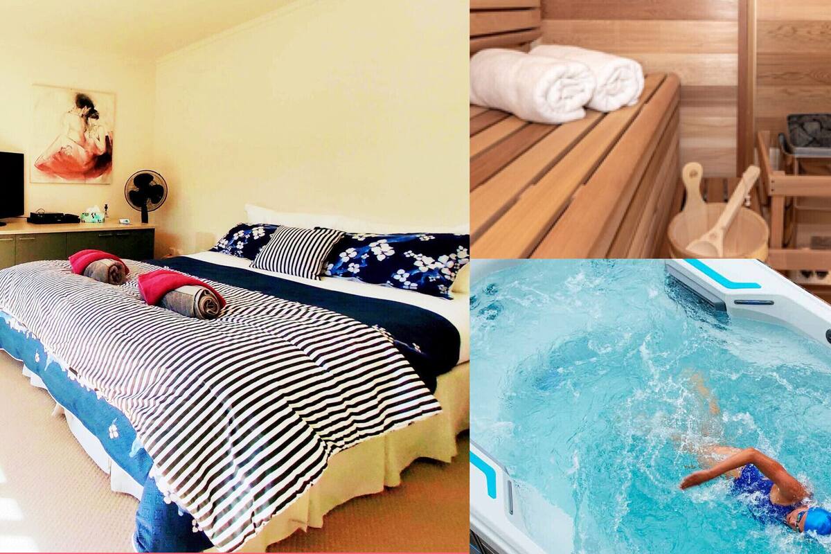 A bedroom with a bed, a bedside table, and a painting. There is also a wooden bench with towels and a fan.