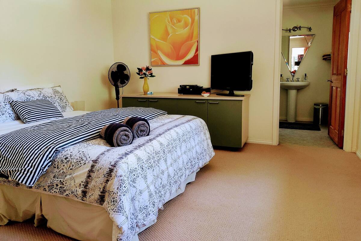 A bedroom with a bed, a desk, a computer, a fan, a vase with flowers, and a bathroom visible through an open door.