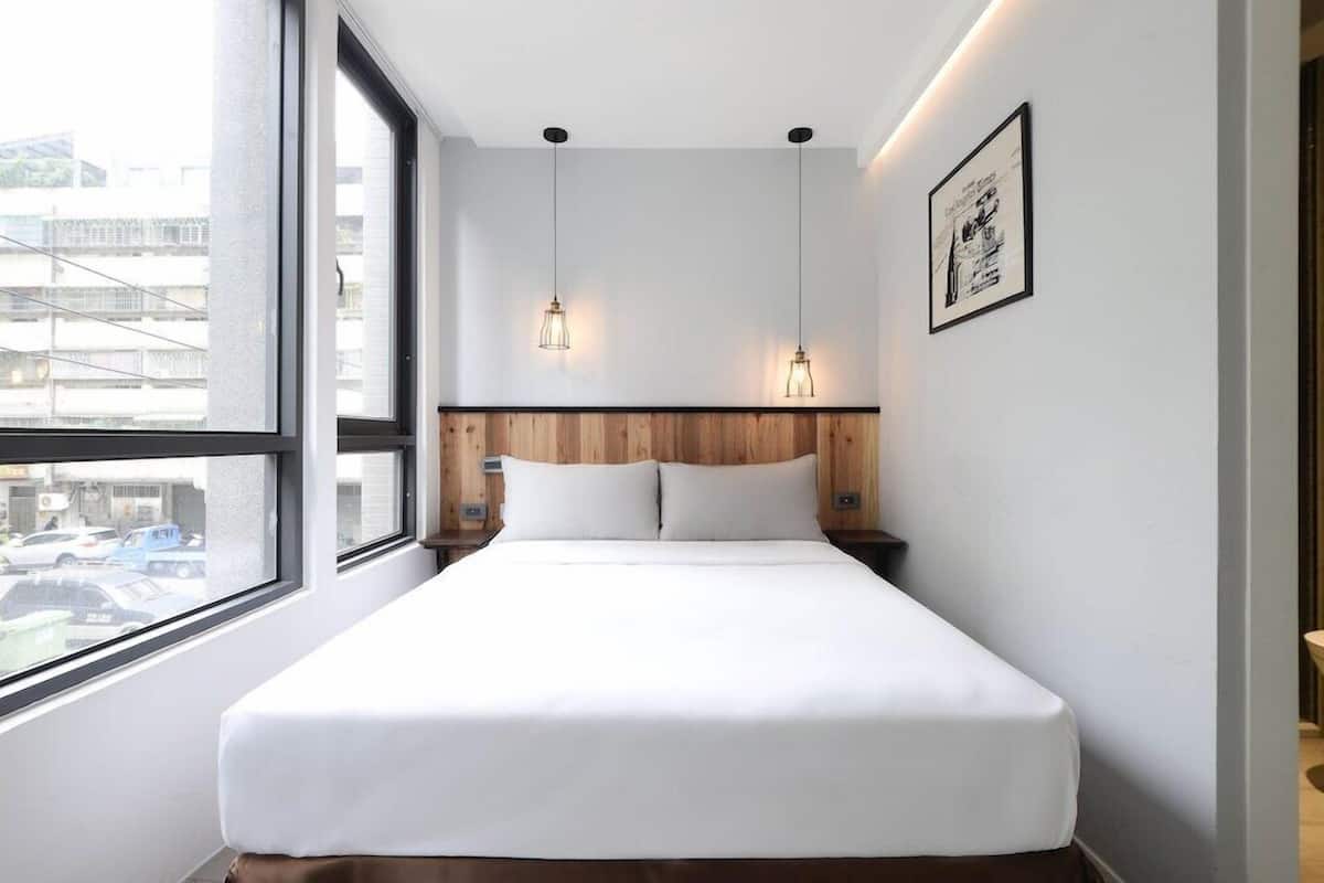 A hotel room with a large window, a bed with white bedding, two pendant lights, and a framed picture on the wall.
