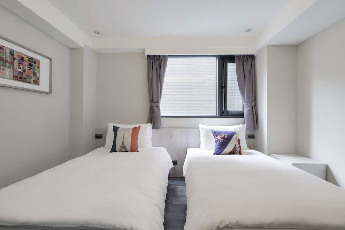 A hotel room with two single beds, a window with curtains, a wall-mounted artwork, and a small bedside table.