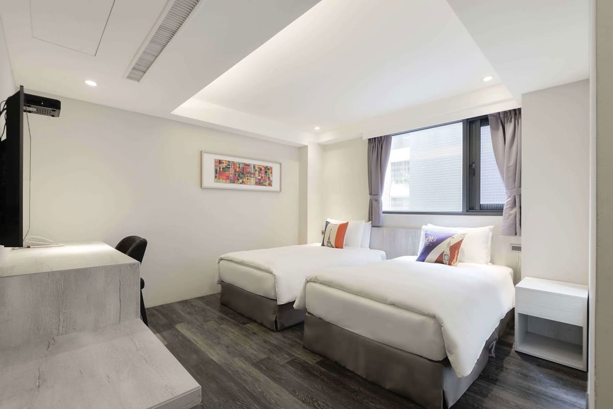 A hotel room with two beds, a desk, a flat-screen TV, and a window with curtains.