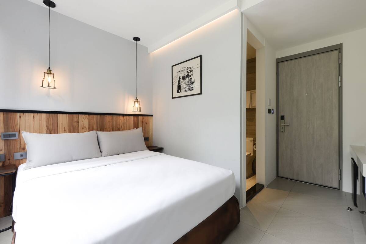 A modern hotel room with a wooden headboard, a large bed, bedside lamps, a framed picture on the wall, and a door leading to the bathroom.