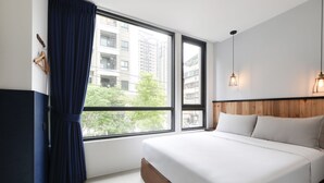 Ein Hotelzimmer mit einem großen Bett, einem Holz-Kopfteil, einem Fenster mit Vorhängen und Blick auf Gebäude und Bäume.