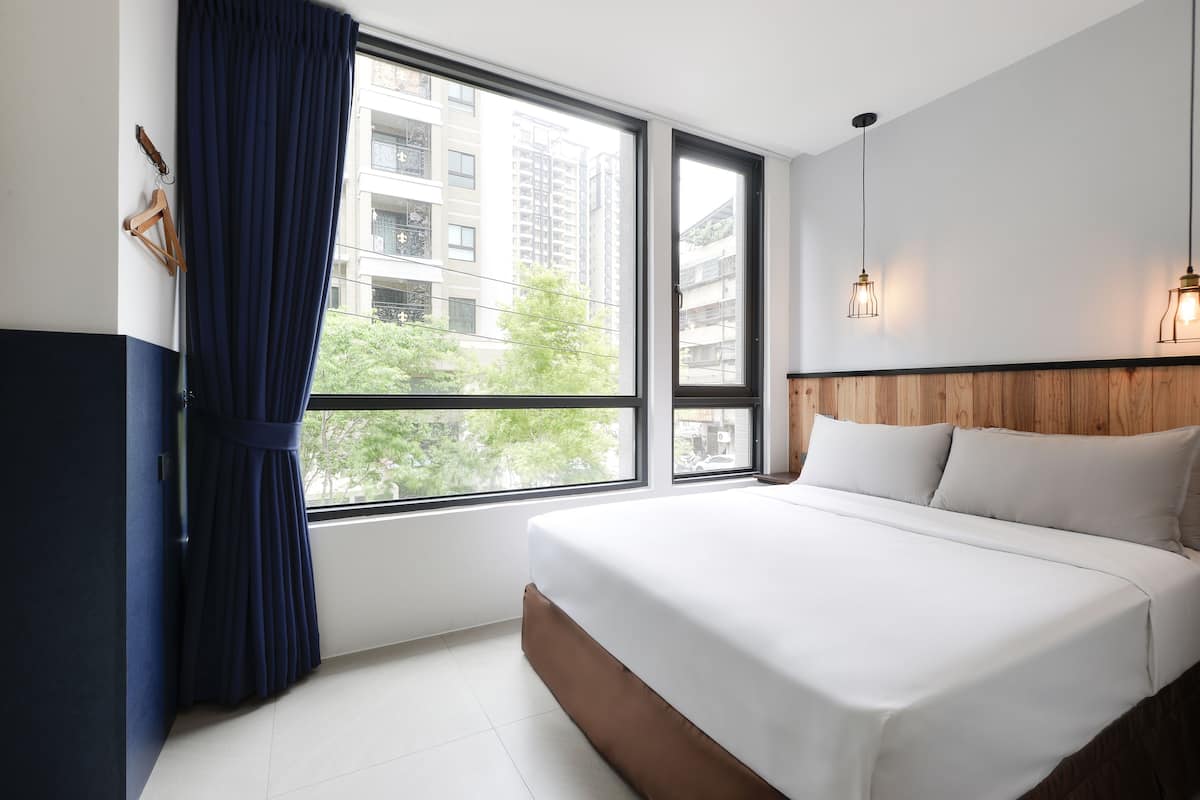 A hotel room with a large bed, a wooden headboard, a window with curtains, and a view of buildings and trees outside.