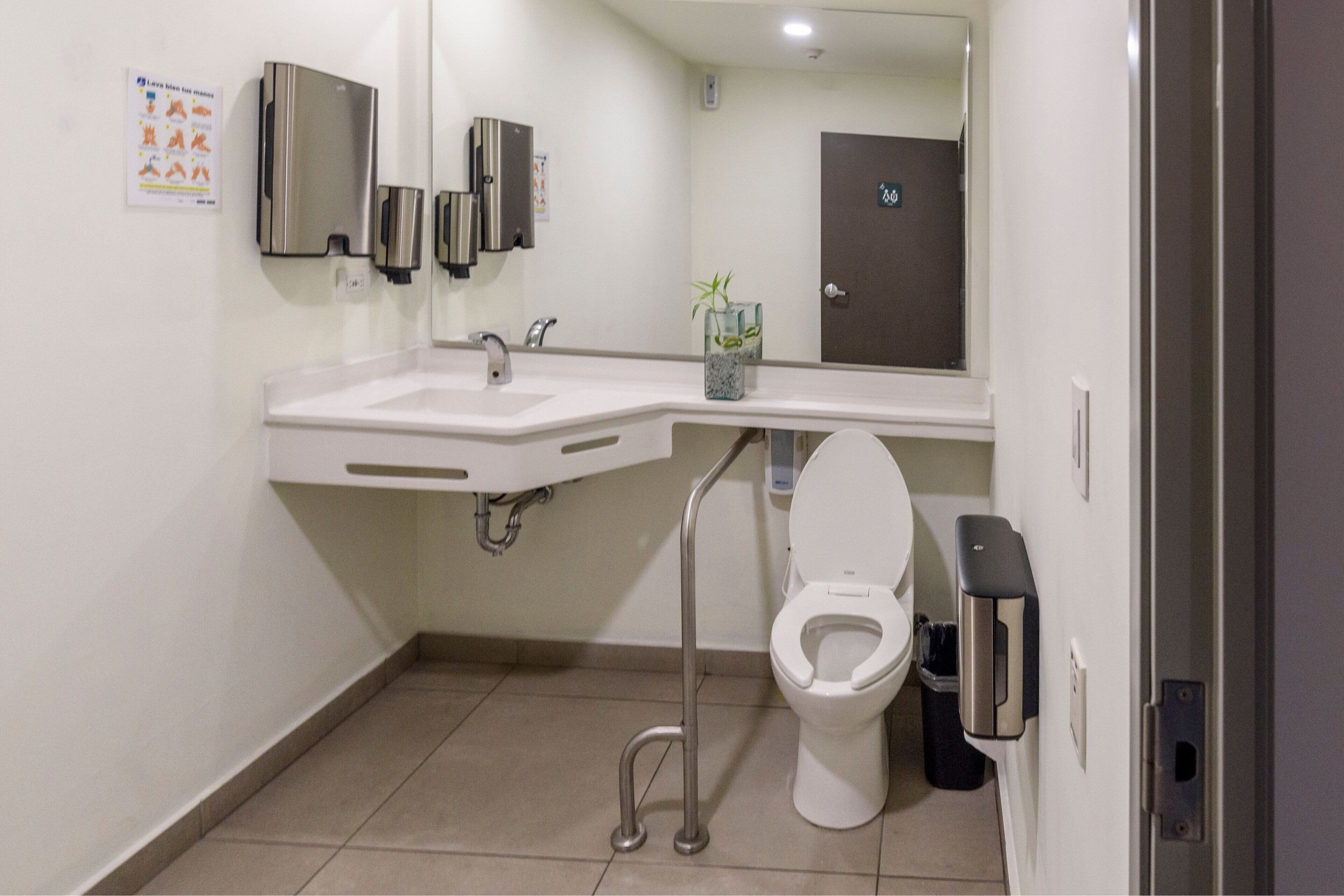 Une salle de bain moderne avec un lavabo blanc, un robinet en acier inoxydable et des toilettes. Un distributeur de gel hydroalcoolique est fixé au mur, et une poubelle se trouve à côté des toilettes.