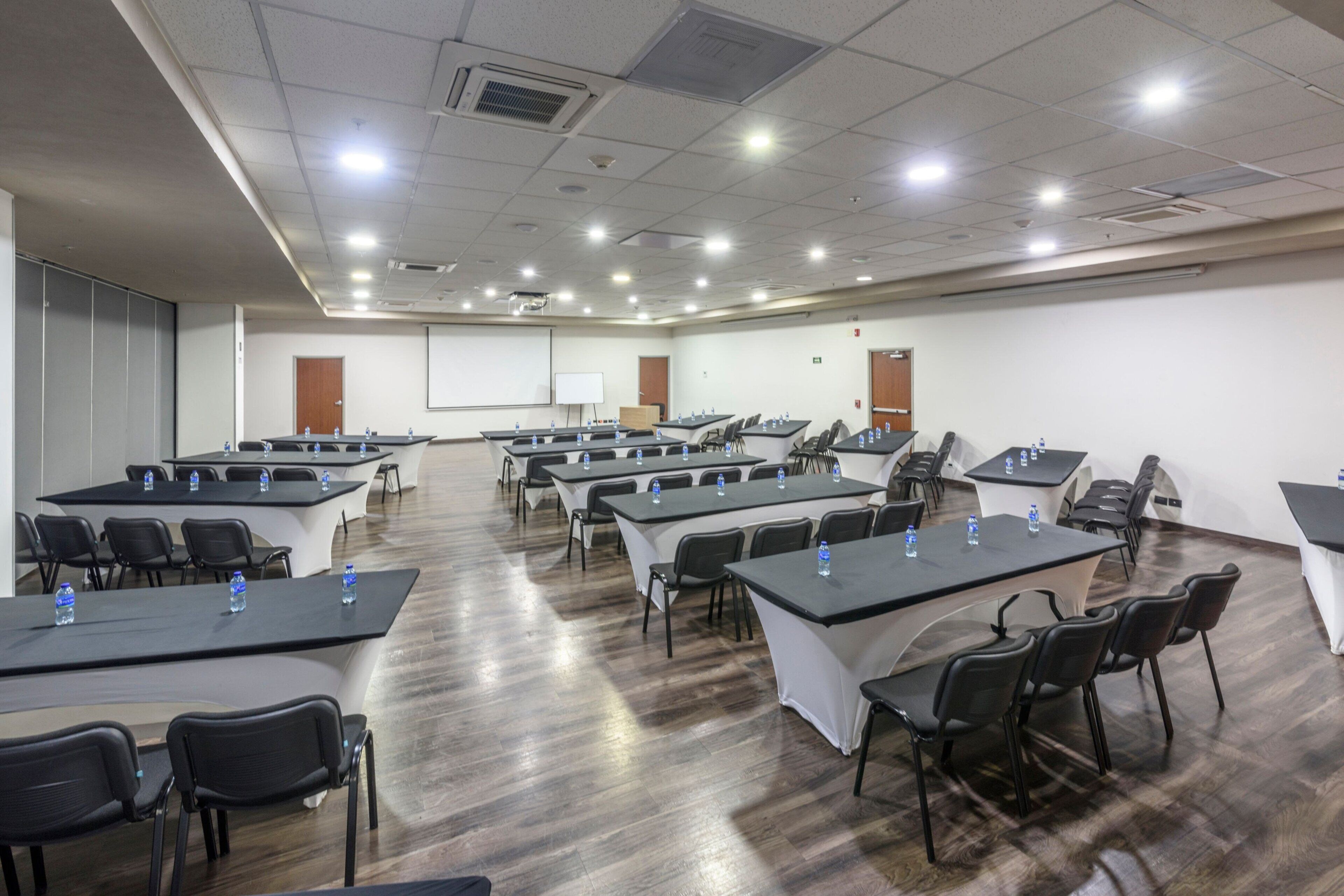 Un salón de conferencias con varias mesas y sillas, equipado con botellas de agua y una pizarra en la pared.