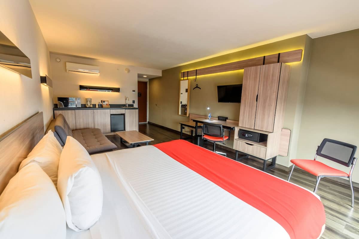 A hotel room with a bed, a sofa, a desk, a chair, a TV, and a kitchenette.