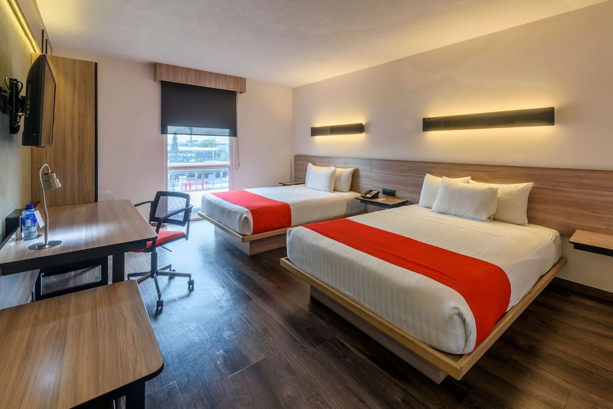 A hotel room with two beds, a desk, and a chair. There is a window with a view of the outside.