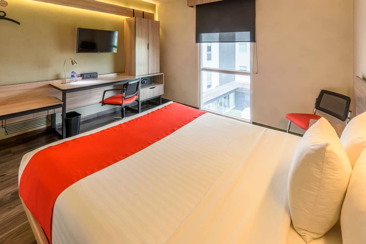 A hotel room with a bed, desk, chair, TV, and a window with a view of buildings.