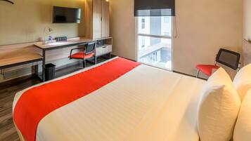 A hotel room with a bed, desk, chair, TV, and a window with a view of buildings.