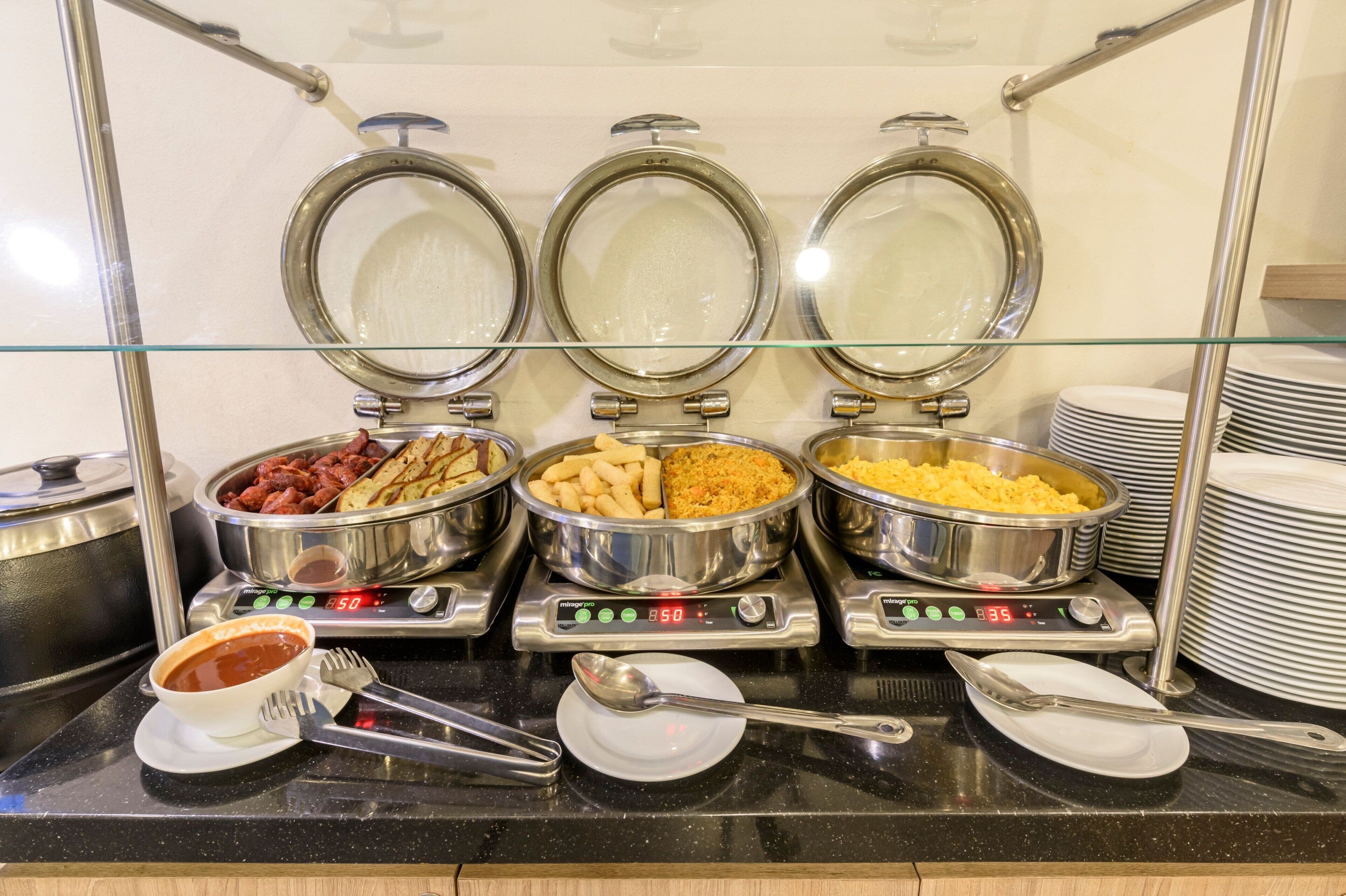 A buffet with three stainless steel chafing dishes containing different foods, a bowl of soup, and a stack of plates.