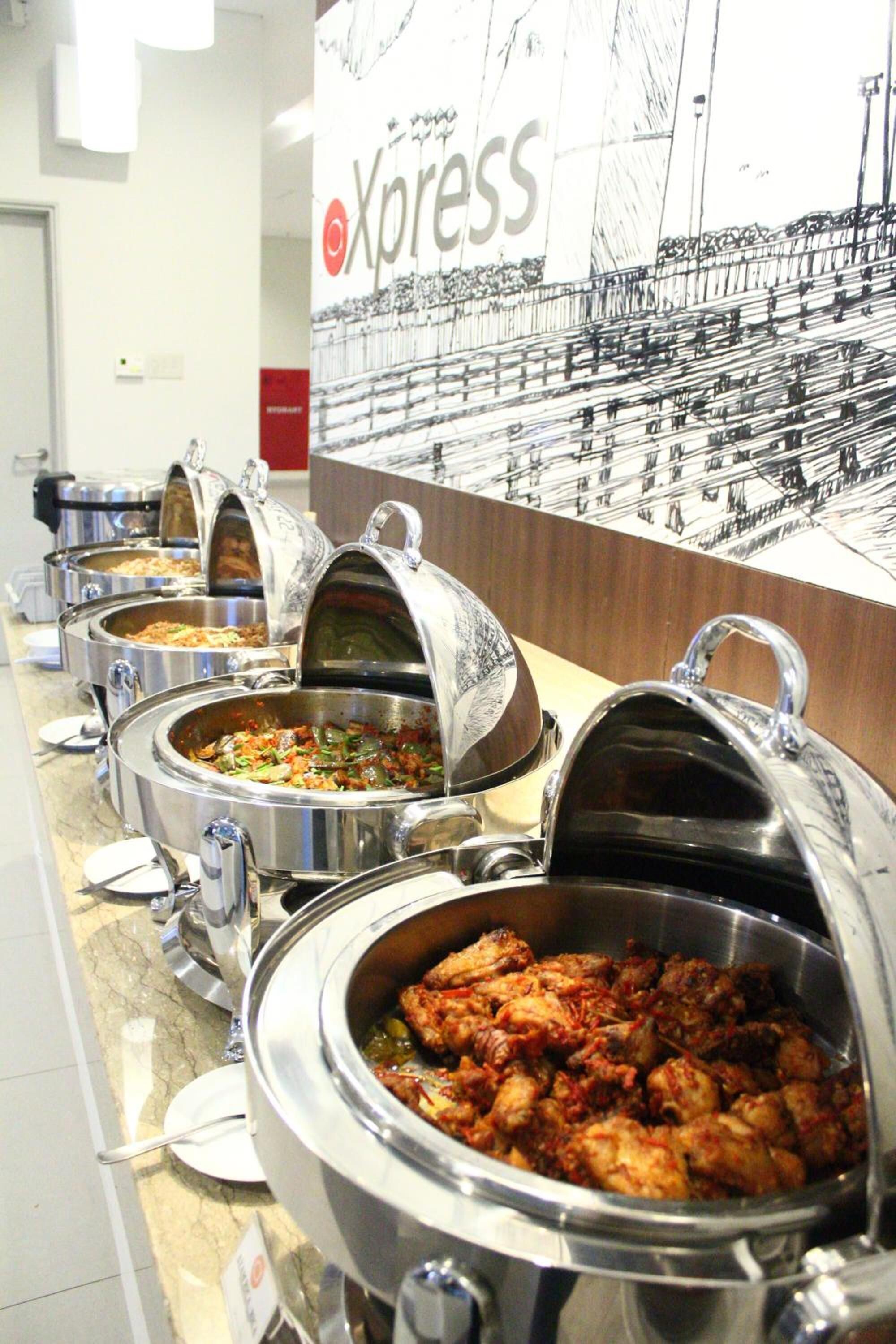 A buffet line with various dishes in metal chafing dishes, a 'Xpress' sign, and a wall with a black and white image.