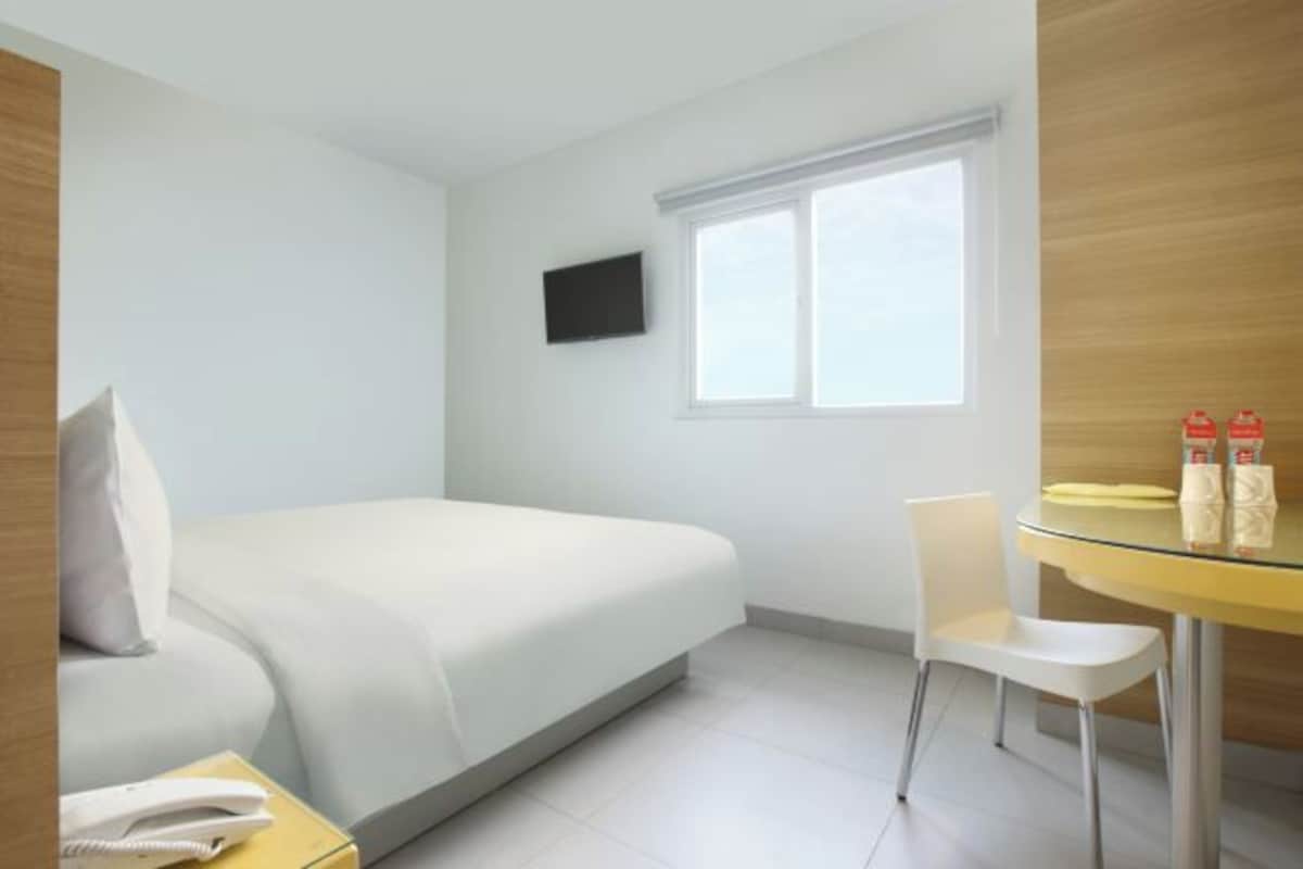 Ein Hotelzimmer mit Bett, einem an der Wand befestigten Fernseher, einem Fenster, einem Sessel und einem kleinen Tisch mit Glasplatte.