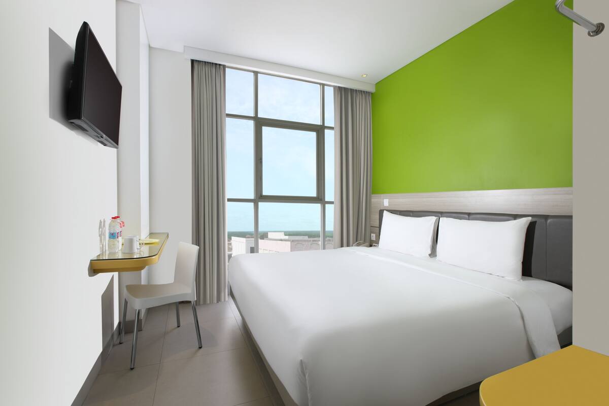 Een hotelkamer met een bed, een bureau met stoel, een televisie en een groot raam met gordijnen.