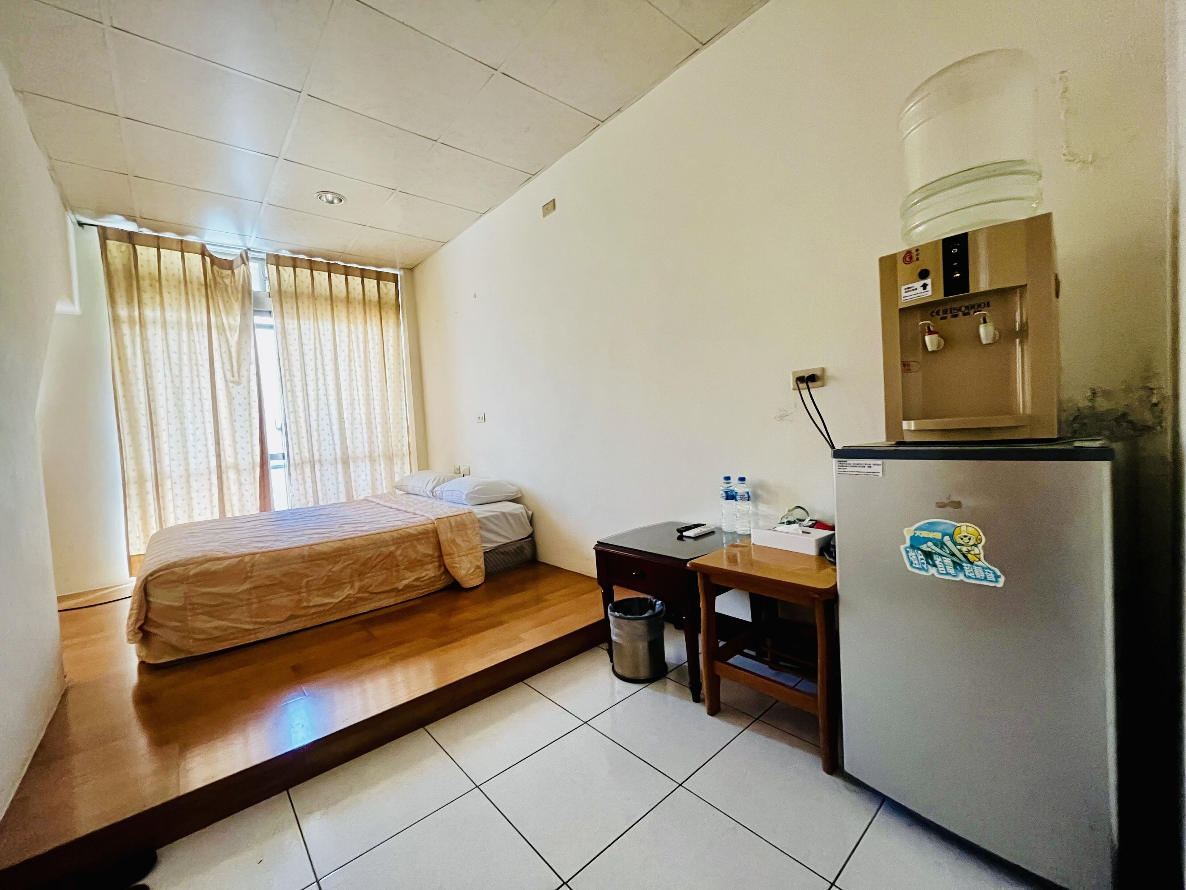 Una habitación de hotel compacta con cama, un escritorio pequeño, un refrigerador y una ventana con cortinas.