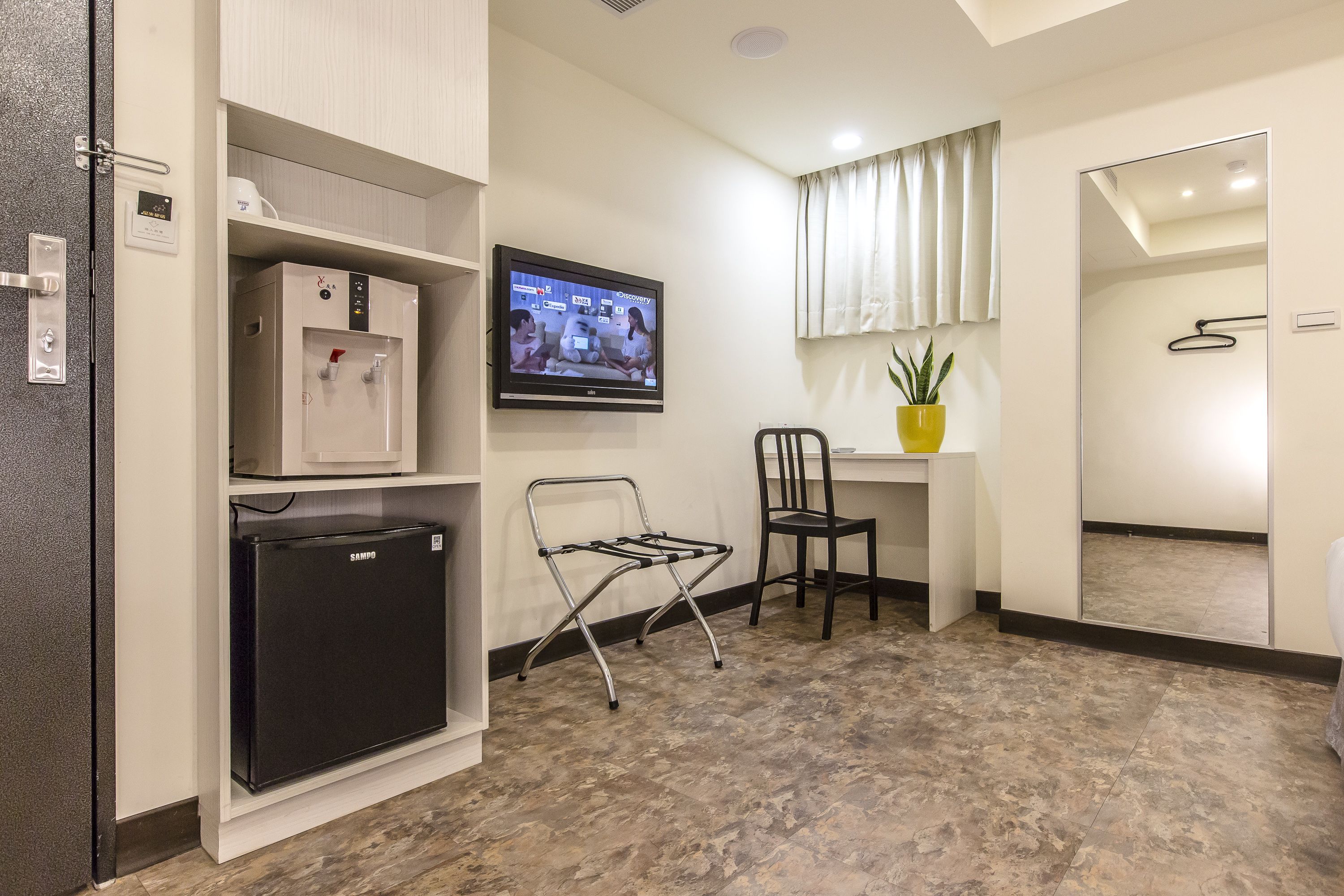 A hotel room with a minibar, a television, a chair, a small table with a plant, and a mirror.