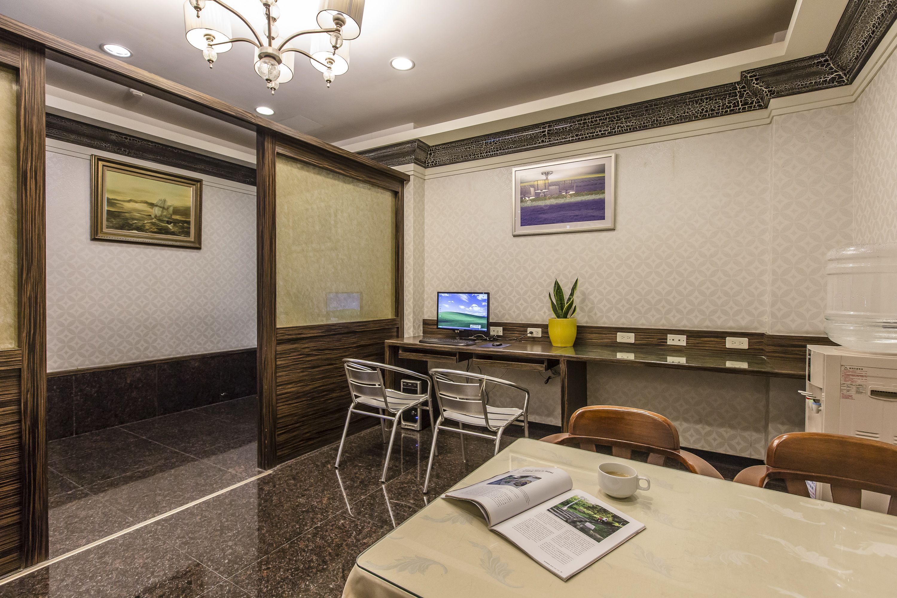 A conference room with a marble floor, a table with a magazine, a coffee cup, and two chairs. There is a computer on a desk with a plant and a water dispenser.
