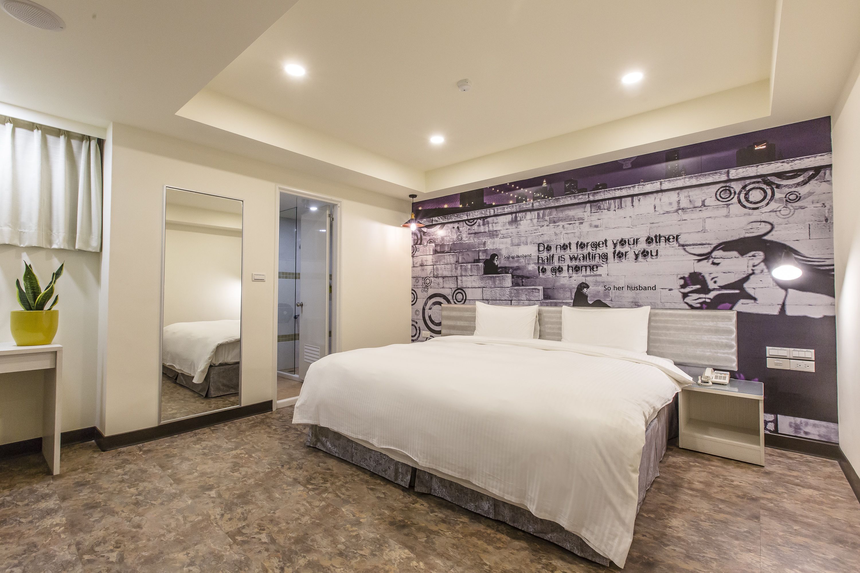 A modern bedroom with a large bed, a mirror, a bedside table, and a wall with a motivational quote.