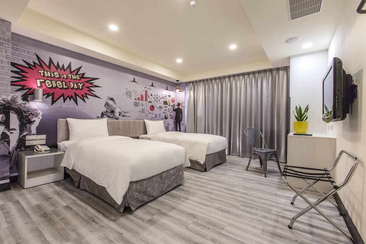 Habitació d'hotel amb dos llits, un escriptori, una cadira, un televisor i una paret amb un mural.