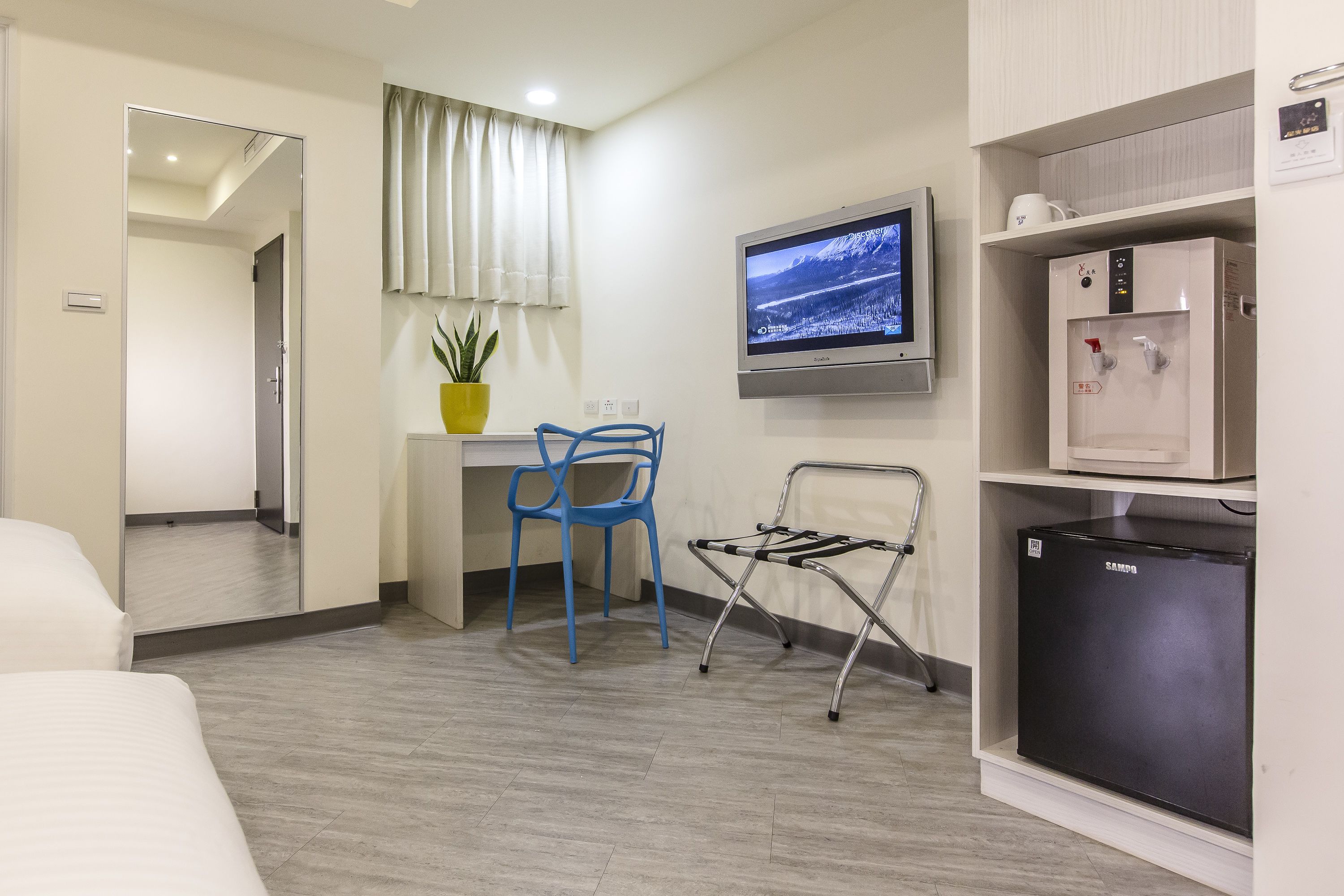 Una habitación de hotel con una cama, un escritorio con una silla, un televisor, un dispensador de agua y un refrigerador.