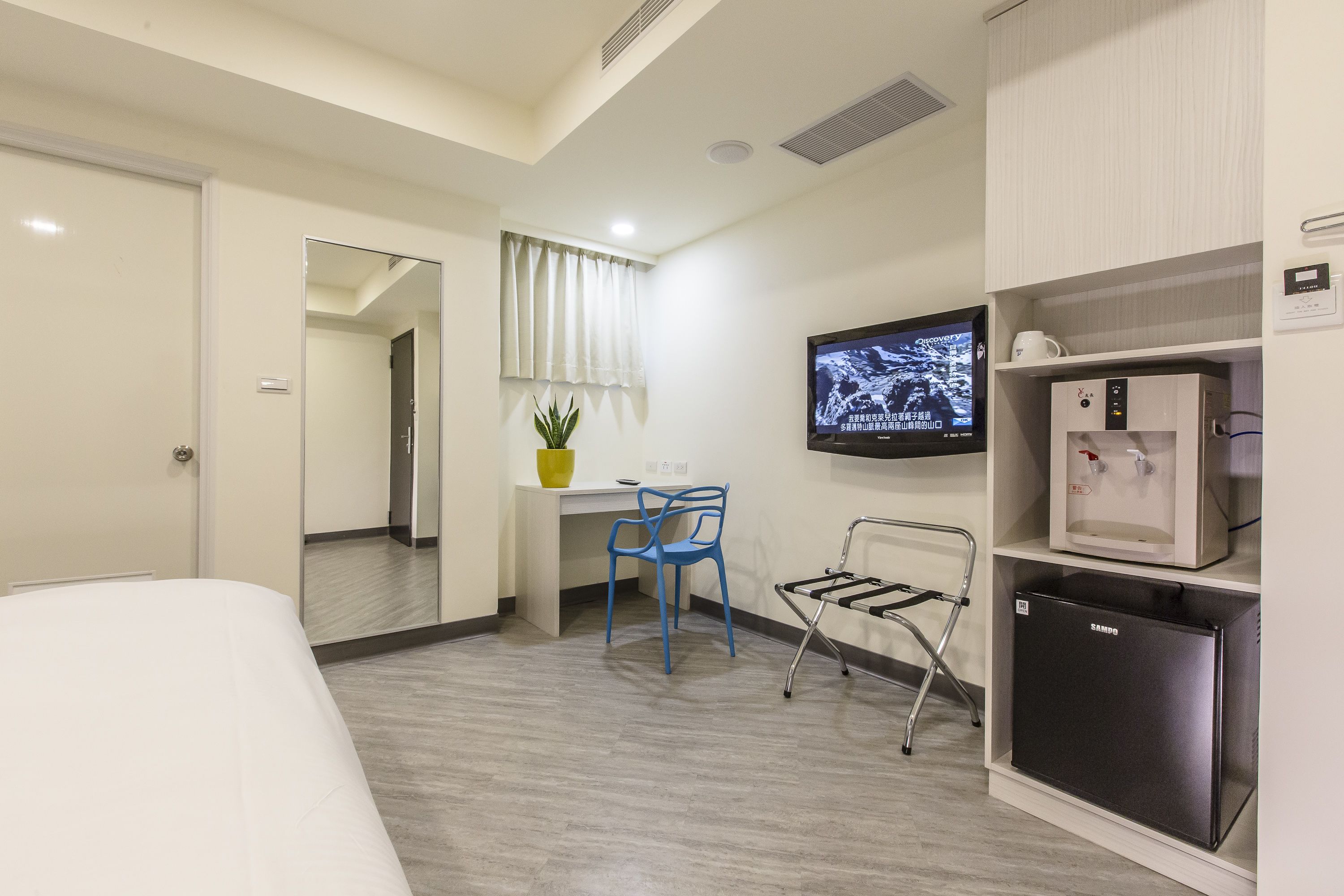 Habitación de hotel con cama, escritorio con silla, televisor, mini refrigerador y dispensador de agua.