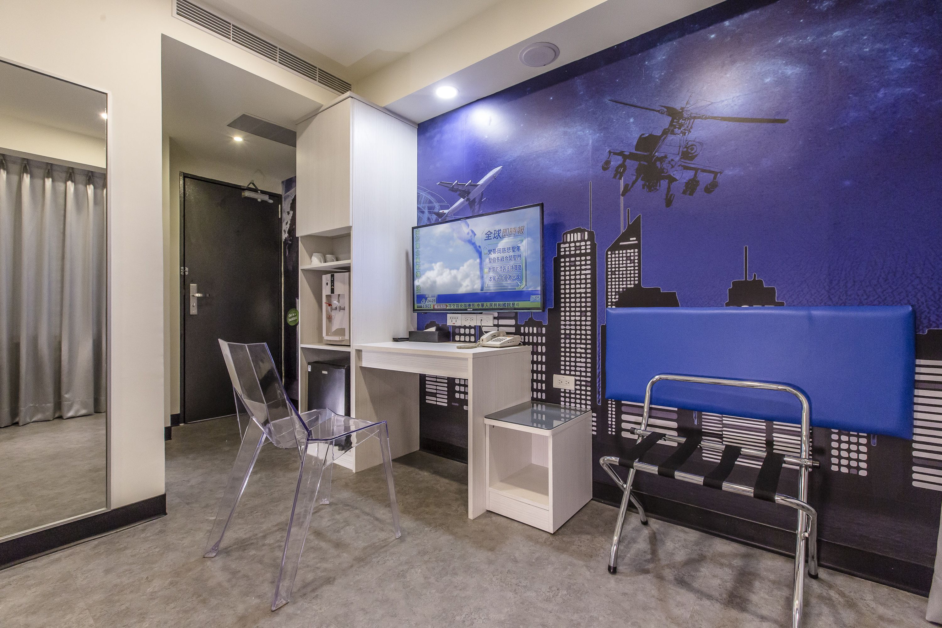 Habitación de hotel moderna con un mural de paisaje urbano, un escritorio con una computadora, una silla y una pequeña mesa.