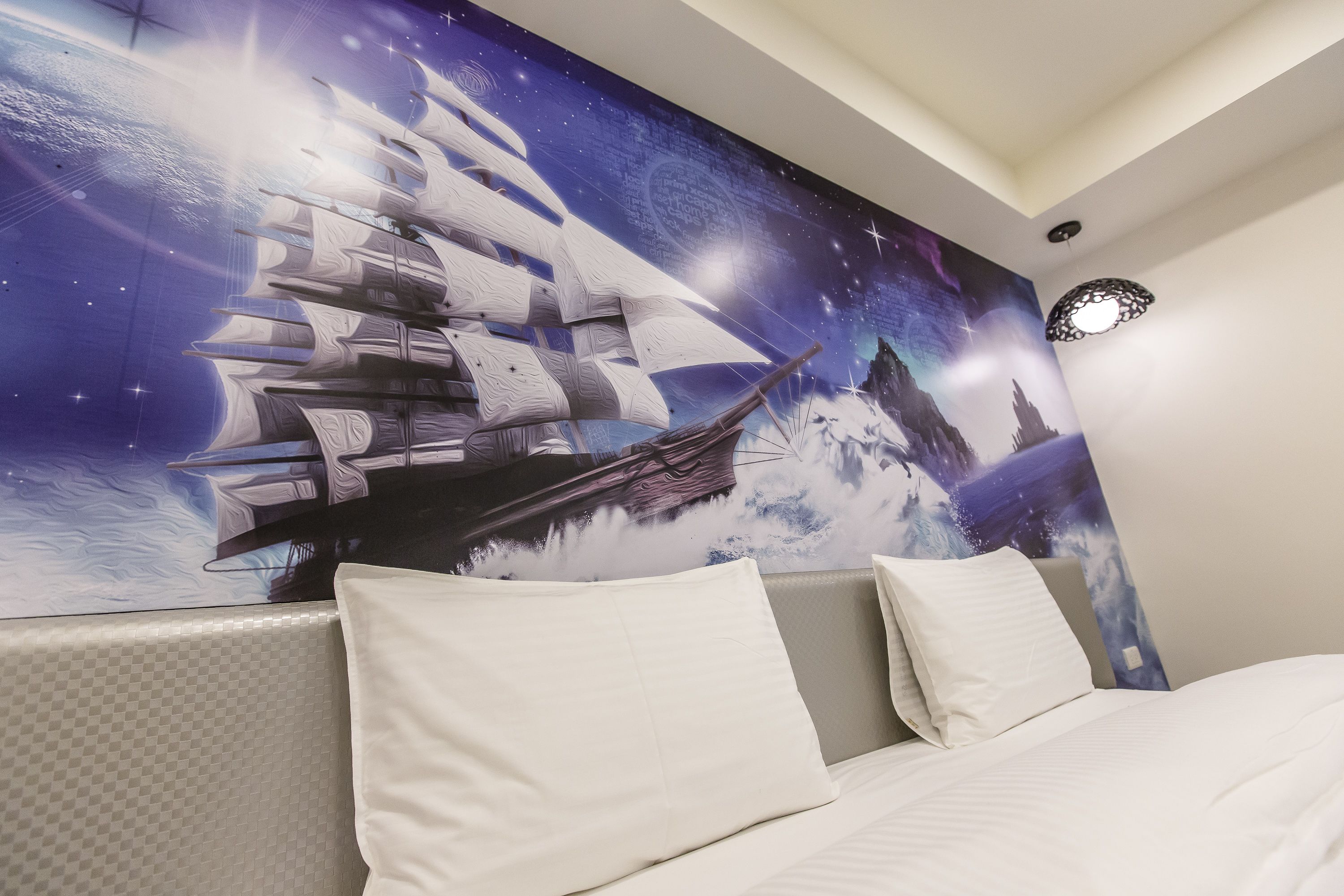 A bedroom with a large wall mural of a ship sailing on water, a bed with white bedding, and a modern ceiling light fixture.