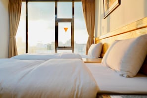 A hotel room with a large bed, a view of the city through a window, and a framed picture on the wall.