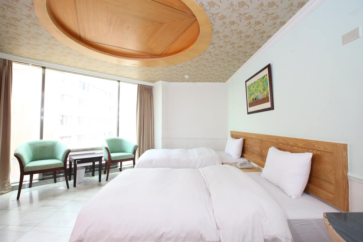Hotellihuone, jossa on kaksi sänkyä, puinen katto, suuri ikkuna, vihreä tuoli ja pieni pöytä.