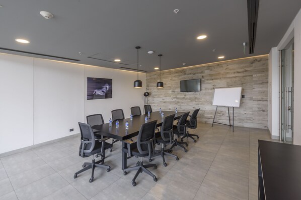 Una sala de conferencias con una mesa larga, sillas negras, una pizarra y un televisor montado en una pared de madera.