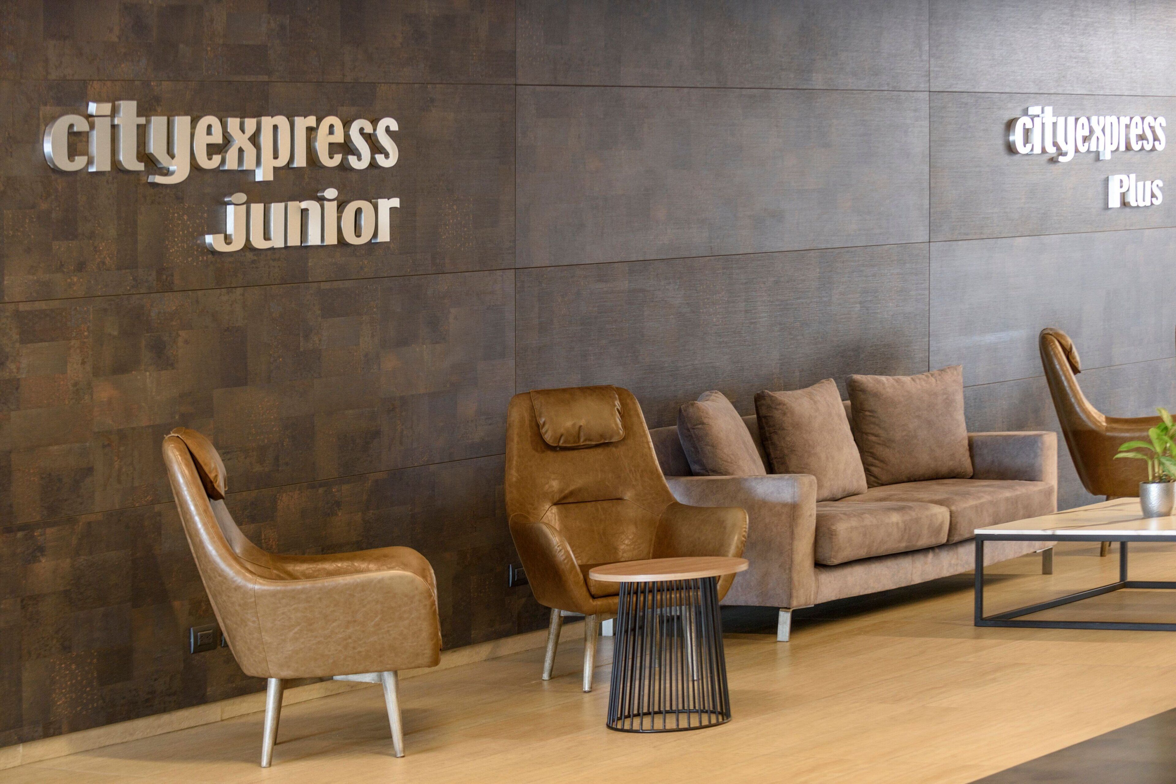 Un área de descanso moderna con un sofá y un sillón de cuero marrón, y una pequeña mesa redonda. En la pared, un letrero indica "cityexpress junior".