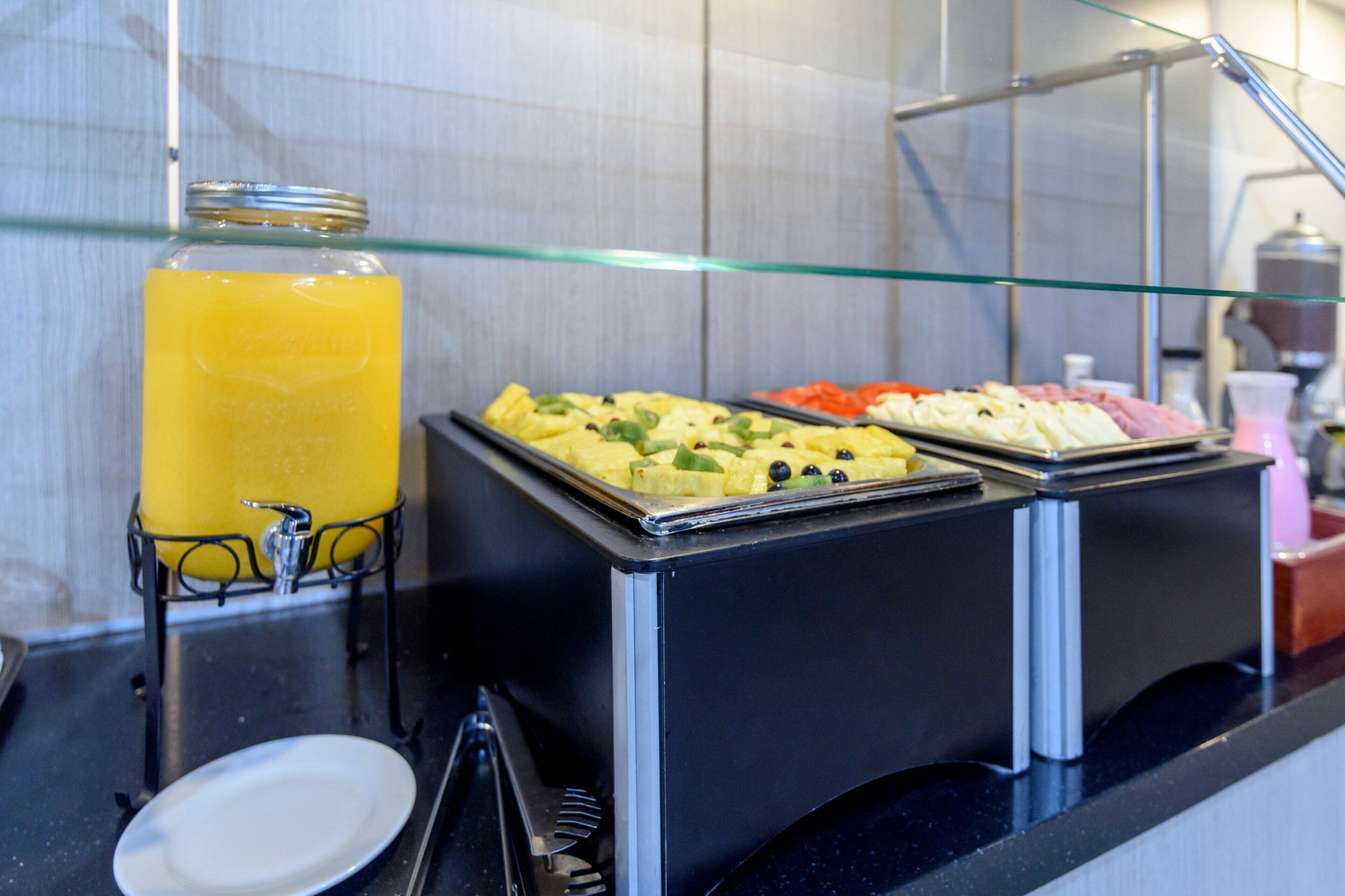 Un bufé con un recipiente de vidrio con jugo de naranja, una bandeja de fruta y una bandeja de pasteles.