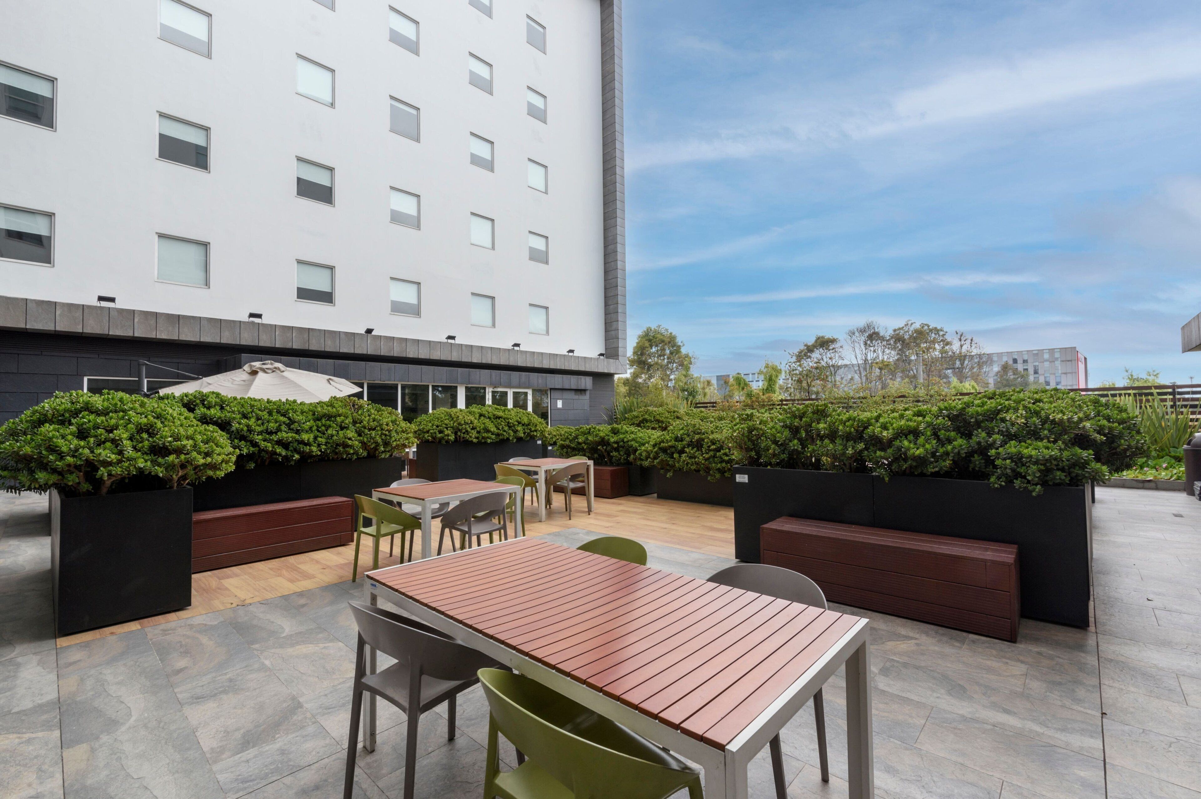 An outdoor patio area with seating arrangements, including tables and chairs, surrounded by greenery and a modern building in the background.