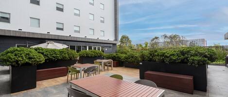 An outdoor patio area with seating arrangements, including tables and chairs, surrounded by greenery and a modern building in the background.