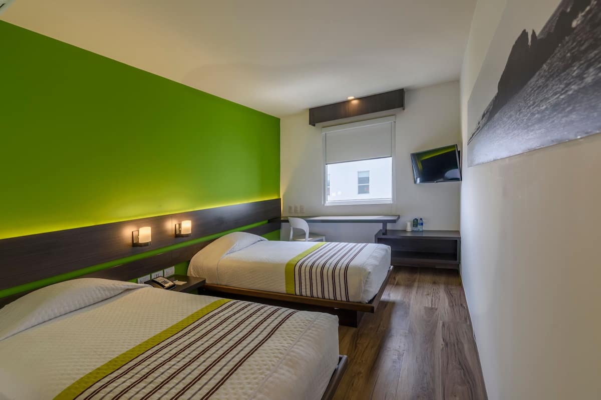 A hotel room with two beds, a green accent wall, a window with a view, a desk with a chair, and a mounted TV.