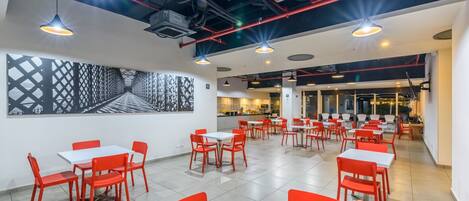 A modern dining area with red chairs and white tables, a kitchen in the background, and a large abstract art piece on the wall.