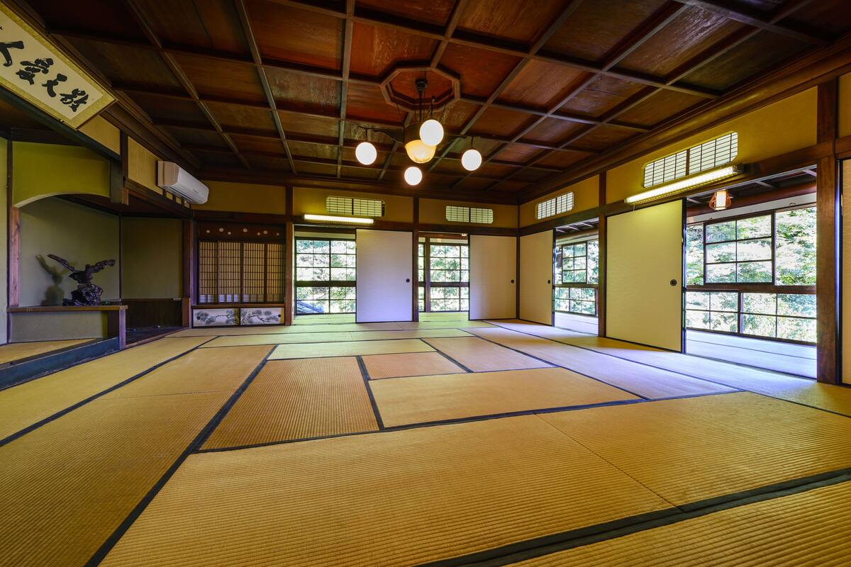A tatami mat room with wooden beams, tatami mats, sliding doors, and a ceiling fan.