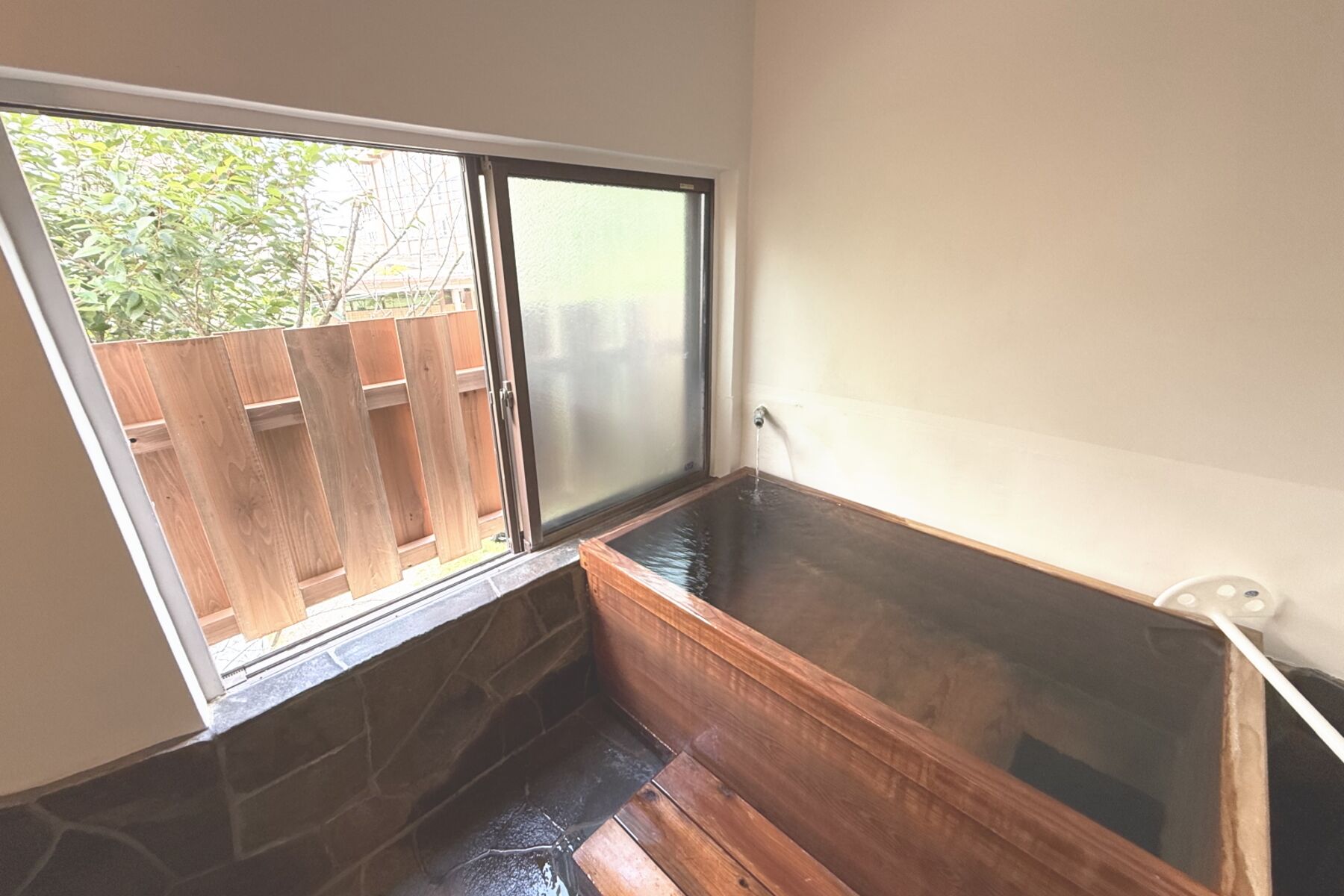 A room with a stone-tiled floor, a wooden bench, a glass shower enclosure, and a view of a wooden fence outside.