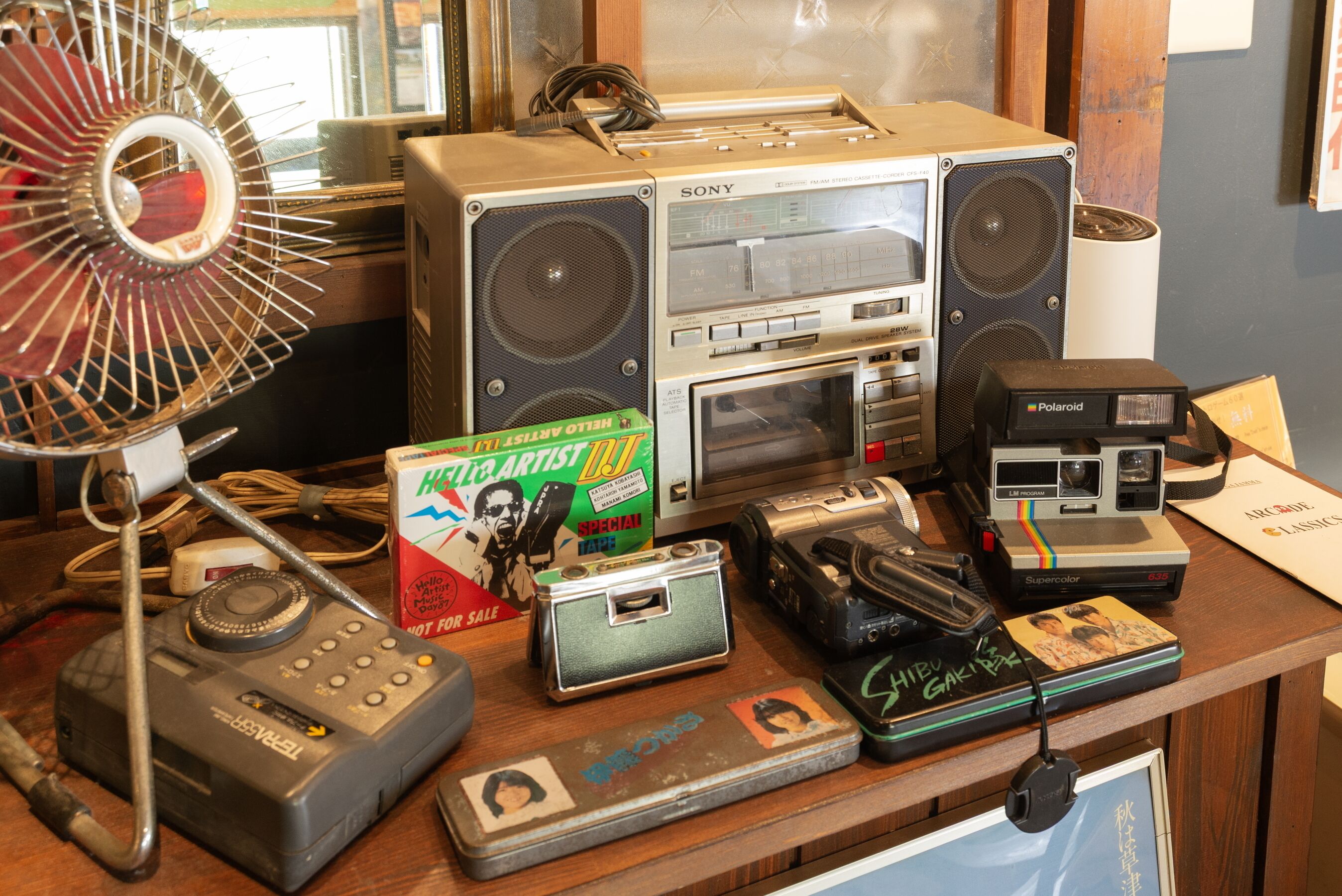 Eine Sammlung von Vintage-Audio-Geräten, darunter ein Sony FM-Stereokassettenrekorder, eine Polaroid-Kamera und ein Ventilator.