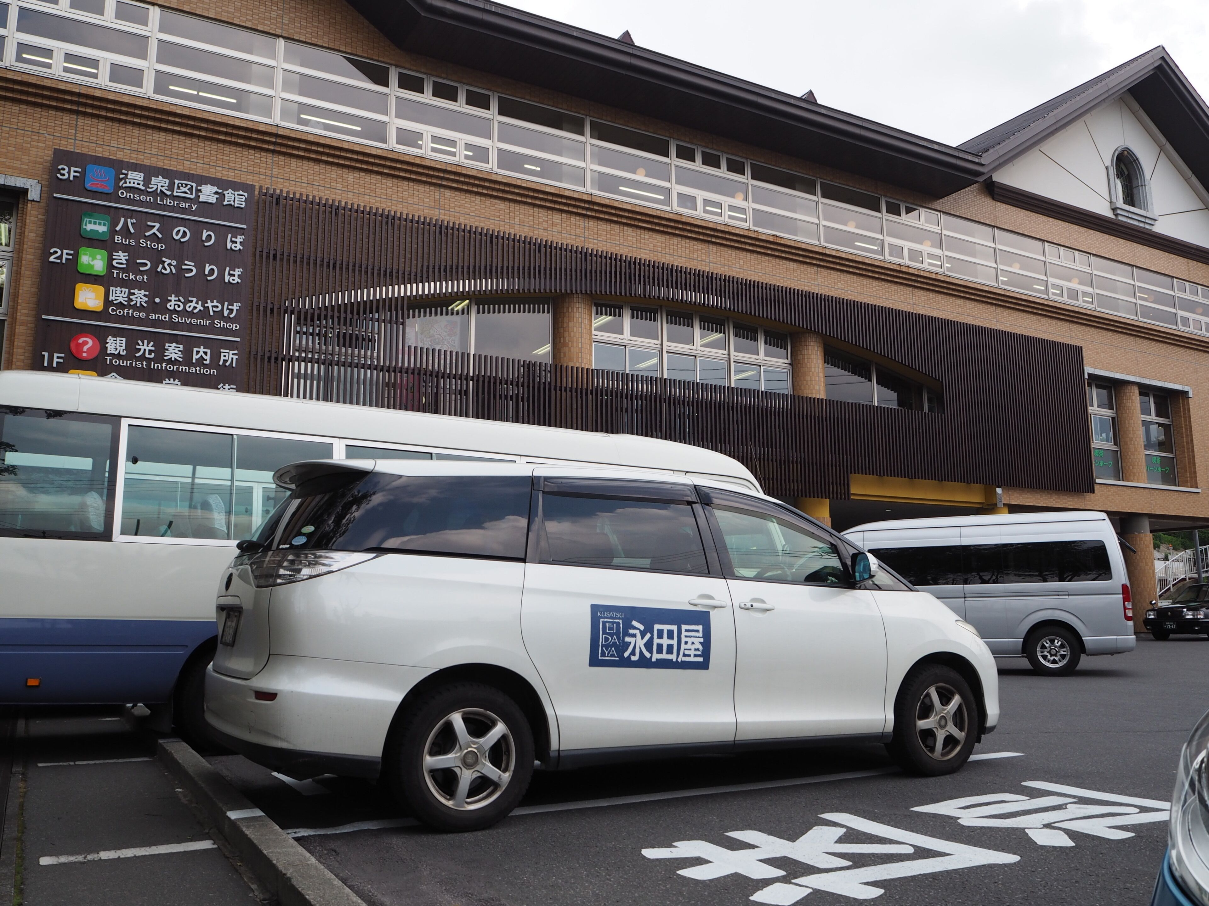Uma área de estacionamento com uma van branca identificada como 'KISSASI ELIDA' e um ponto de ônibus com várias indicações.