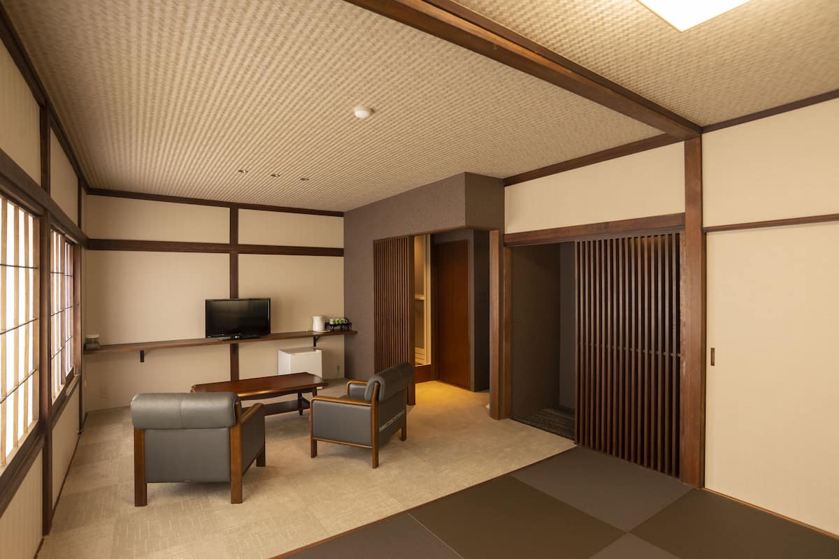 Una habitación japonesa tradicional con suelo de tatami, una mesa baja, dos sillas, un televisor y puertas correderas.