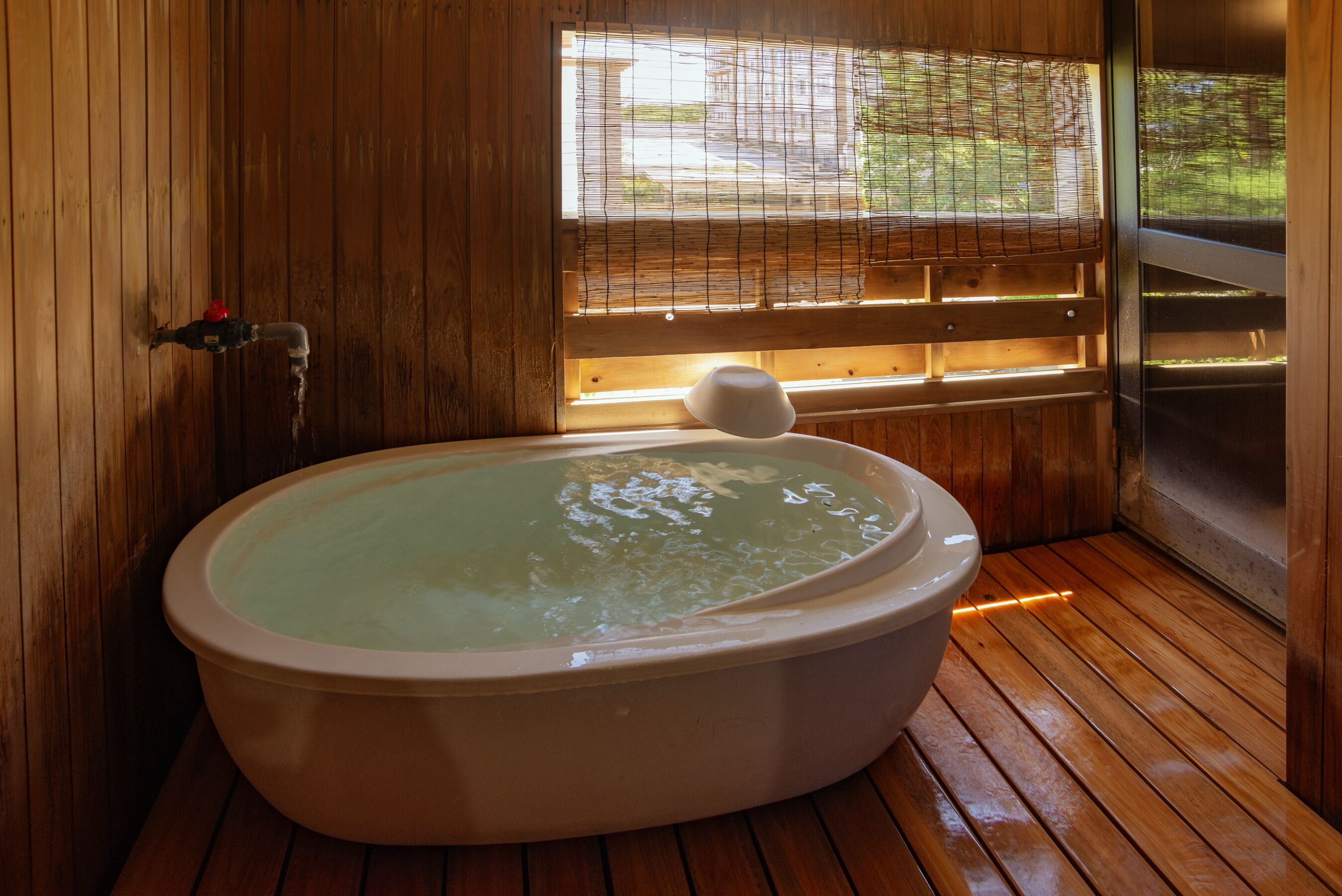 Een badkamer met houten vloer, een vrijstaand bad, een raam met zonwering en een zichtbare waterkraan.