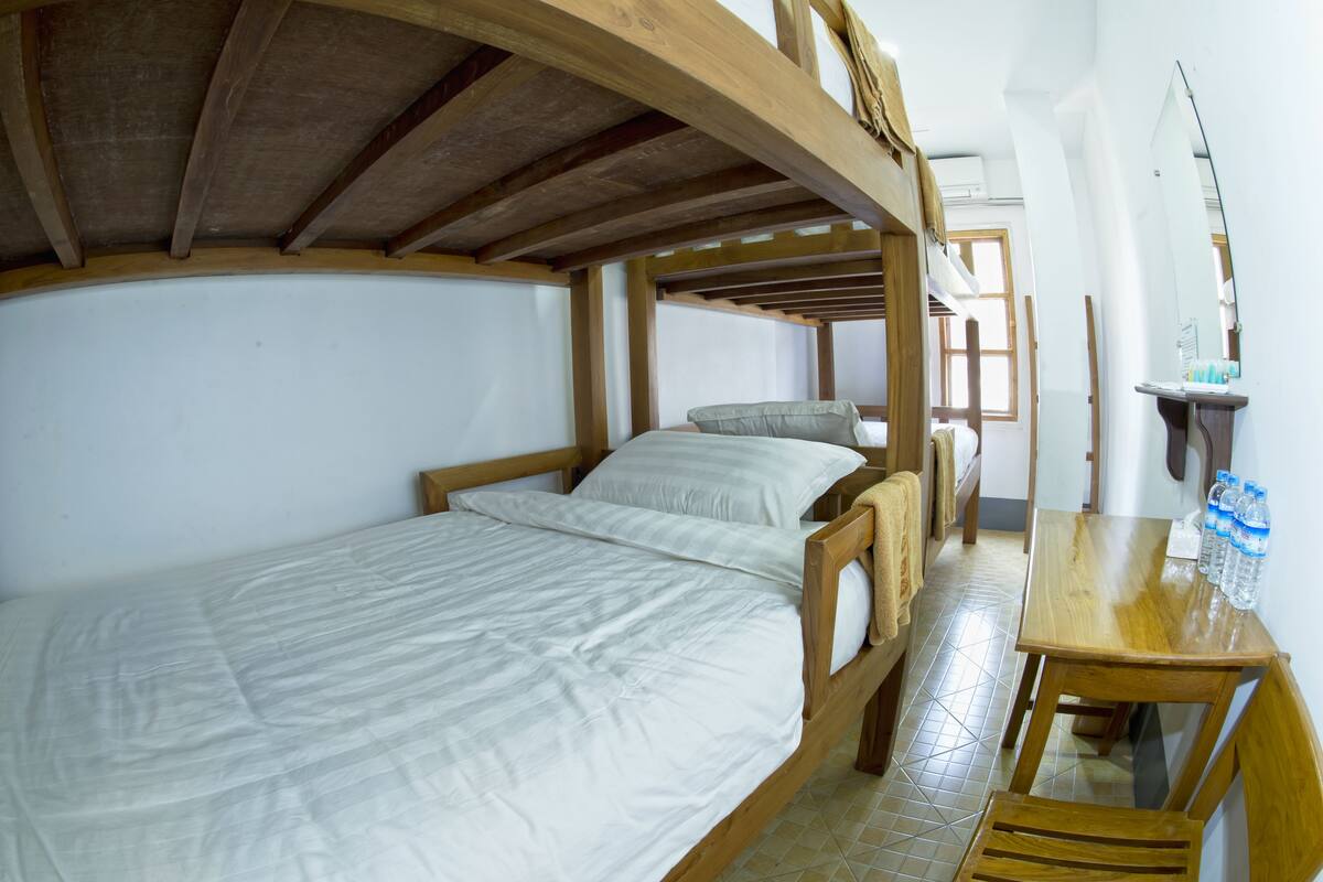 Un lit superposé avec une structure en bois et du linge de lit blanc, une table en bois avec des bouteilles d’eau et un miroir accroché au mur.