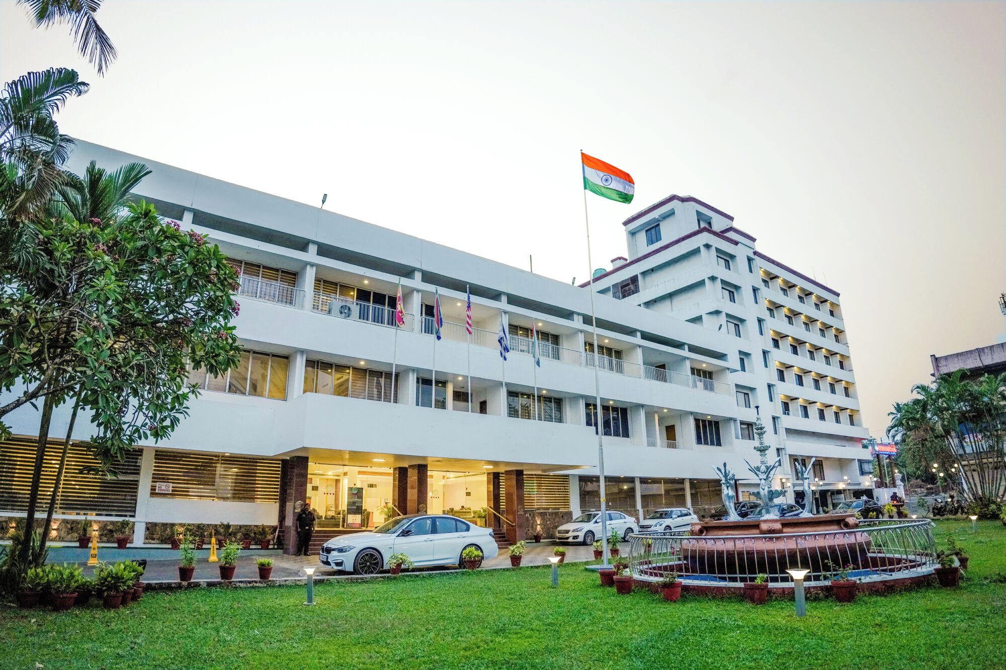 A multi-story hotel with a flag on top, a parking area, and a fountain in the front.