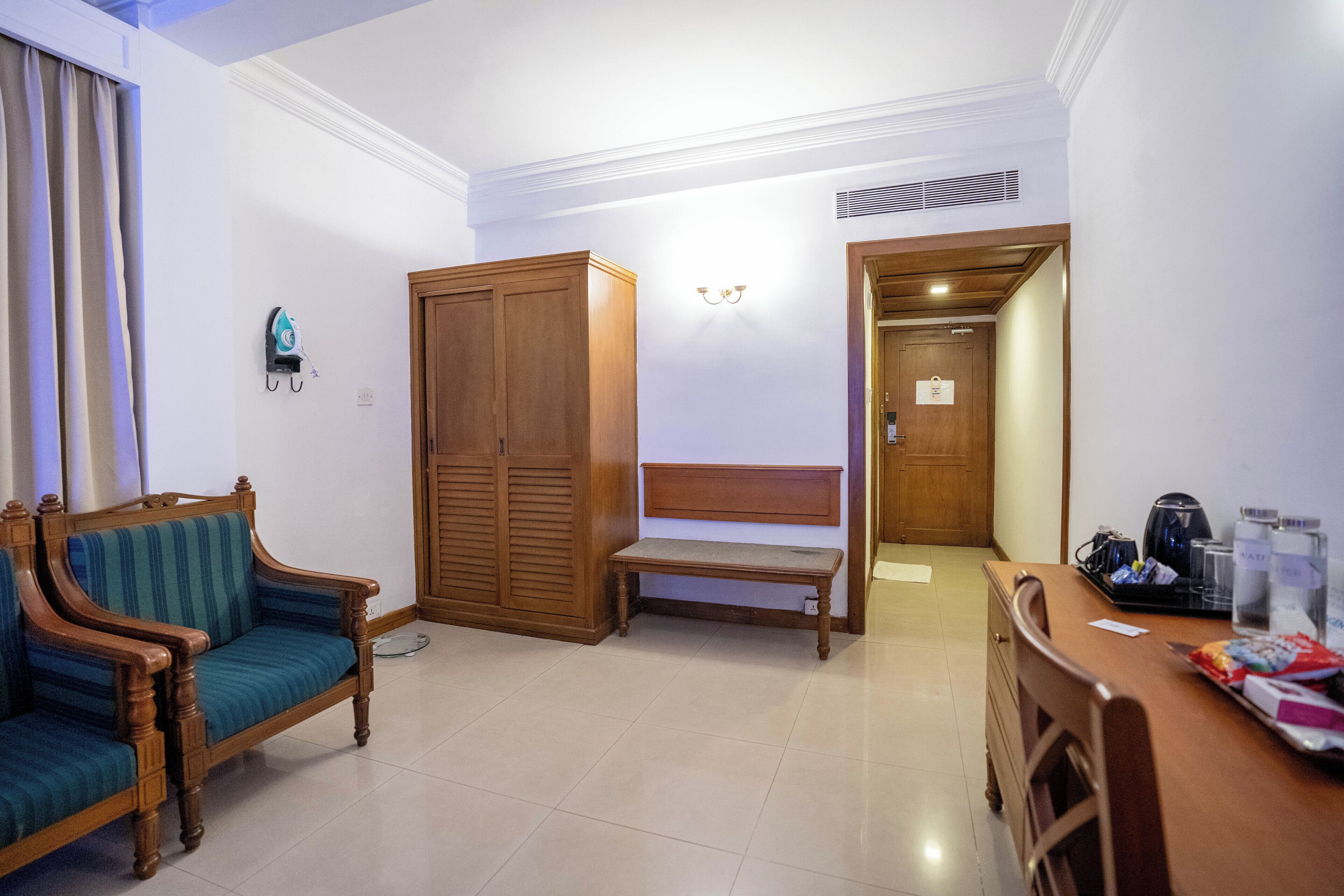 A hotel room with a wooden wardrobe, a bench, a sofa, a dining table, and a kettle.