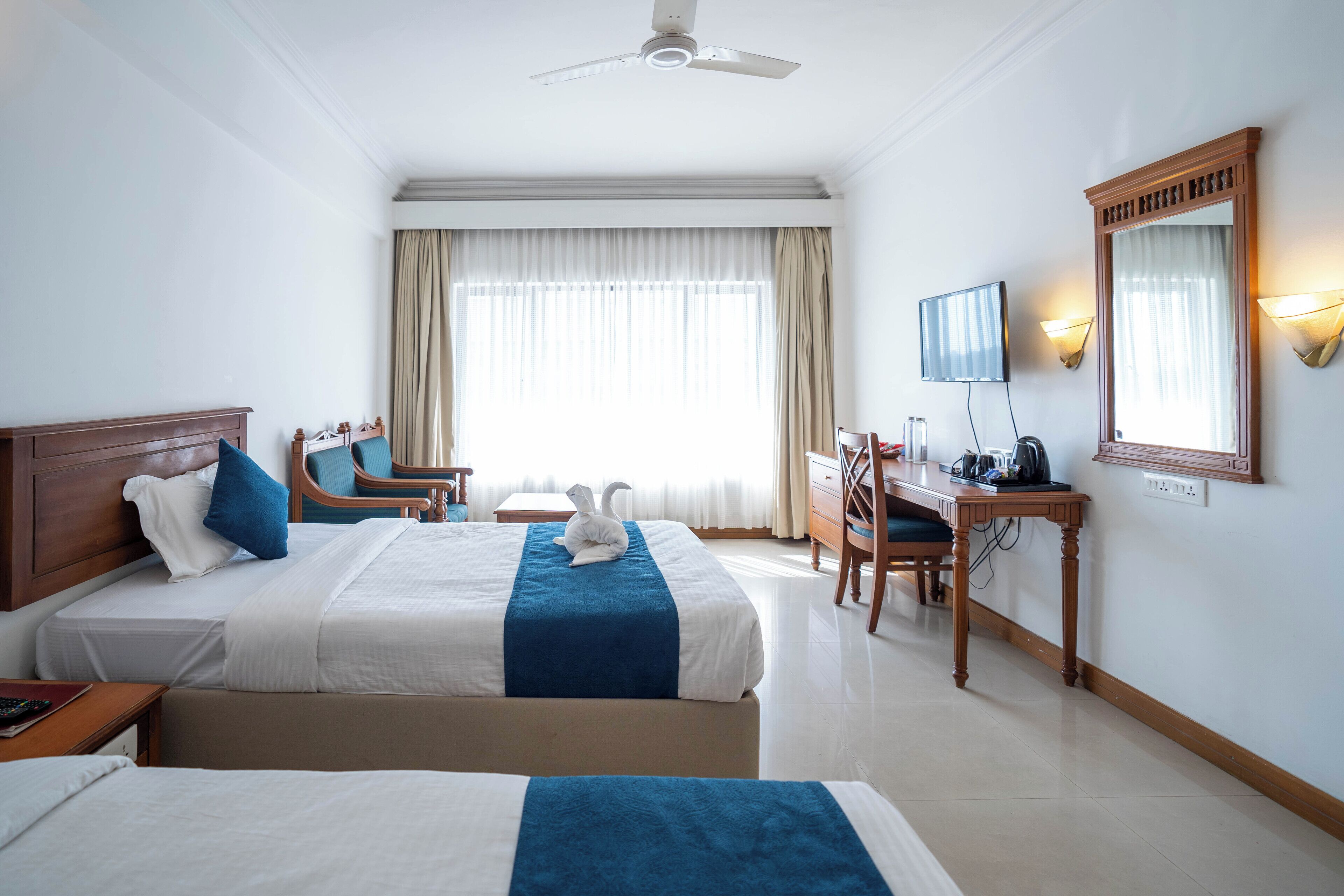 Habitación de hotel con dos camas, un escritorio, una silla, televisión y un ventanal con cortinas.