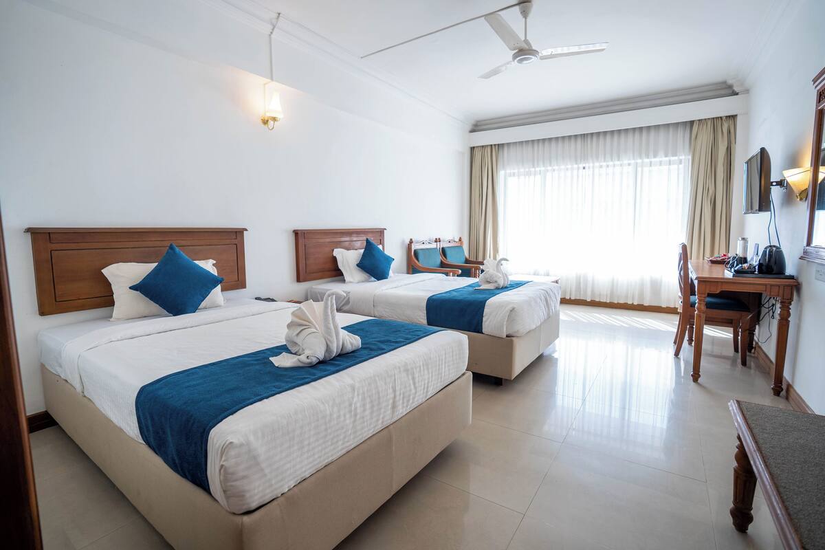 Une chambre d’hôtel avec deux lits, un bureau, une chaise, un ventilateur de plafond et une grande fenêtre avec des rideaux.