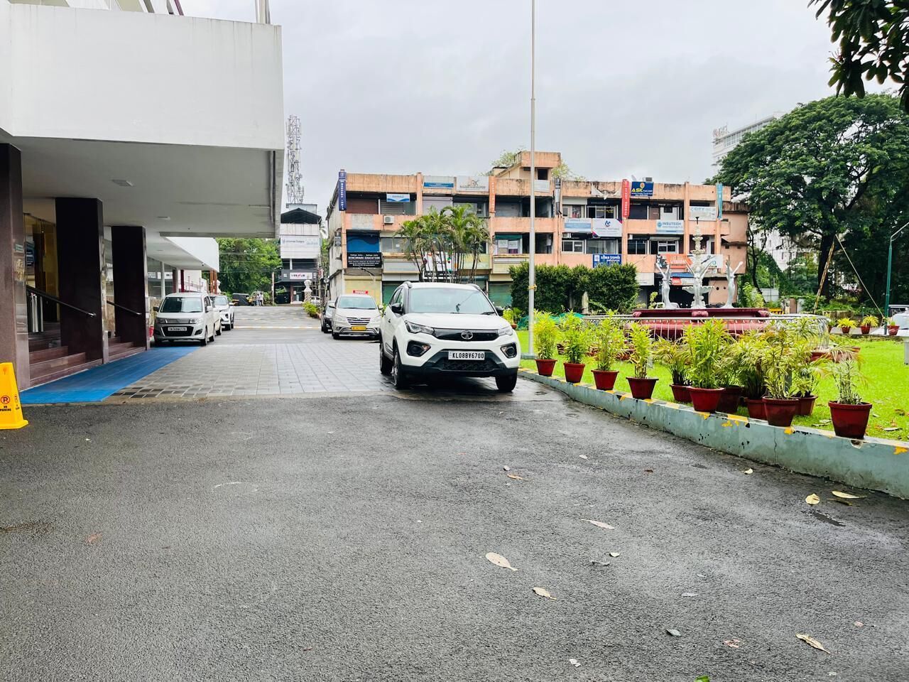 A parking area with multiple cars, a building with a blue entrance, and a row of potted plants along the sidewalk.