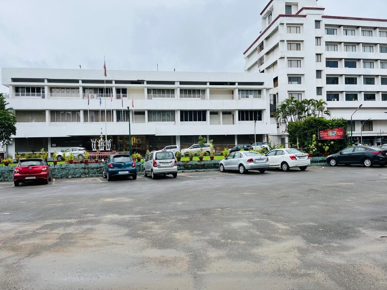 A parking lot with several cars, a building with multiple floors and balconies, and a flag on top.