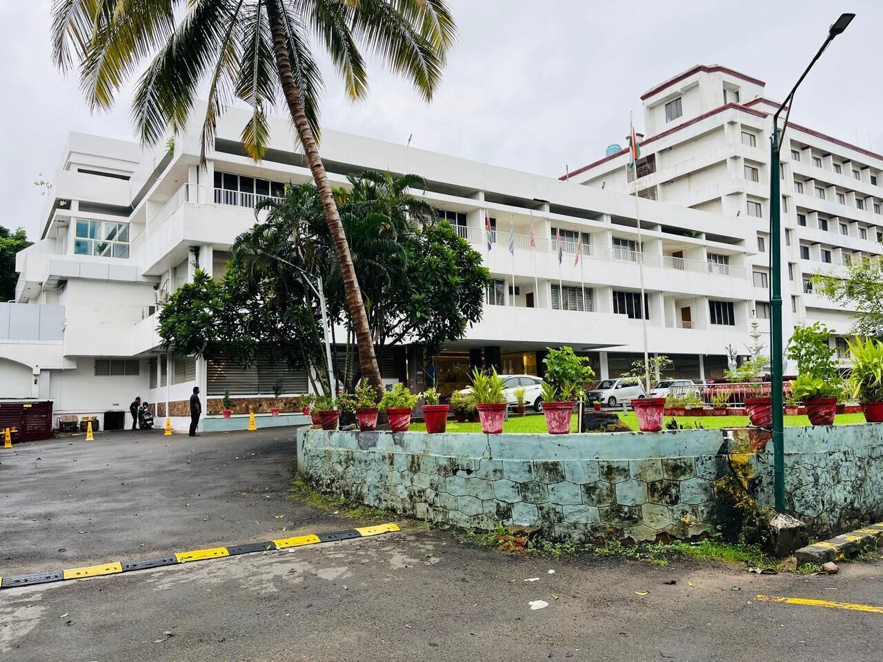 A multi-story building with a white facade, a parking area, and potted plants along the perimeter.
